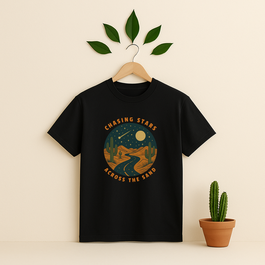 Black t-shirt with 'Chasing Stars Across the Sand' graphic on a wooden hanger against a beige background with a cactus plant.
