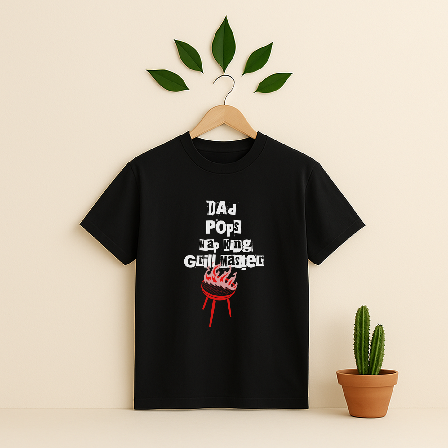 Black t-shirt with 'Dad Pops Grill Master' graphic on a wooden hanger against a beige background with a cactus plant.