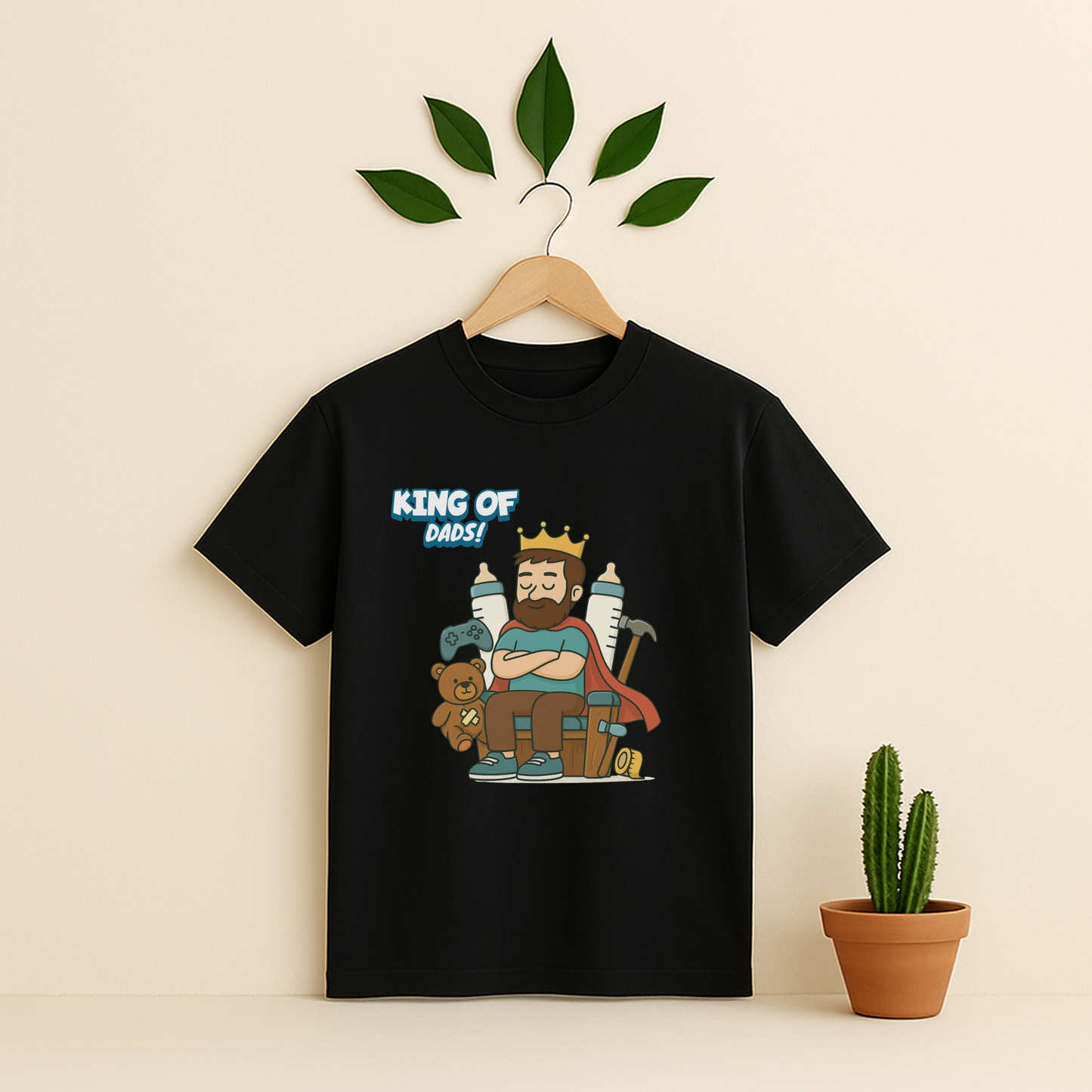 Black t-shirt with 'King Of Dads!' graphic on a wooden hanger against a beige background with a cactus plant.