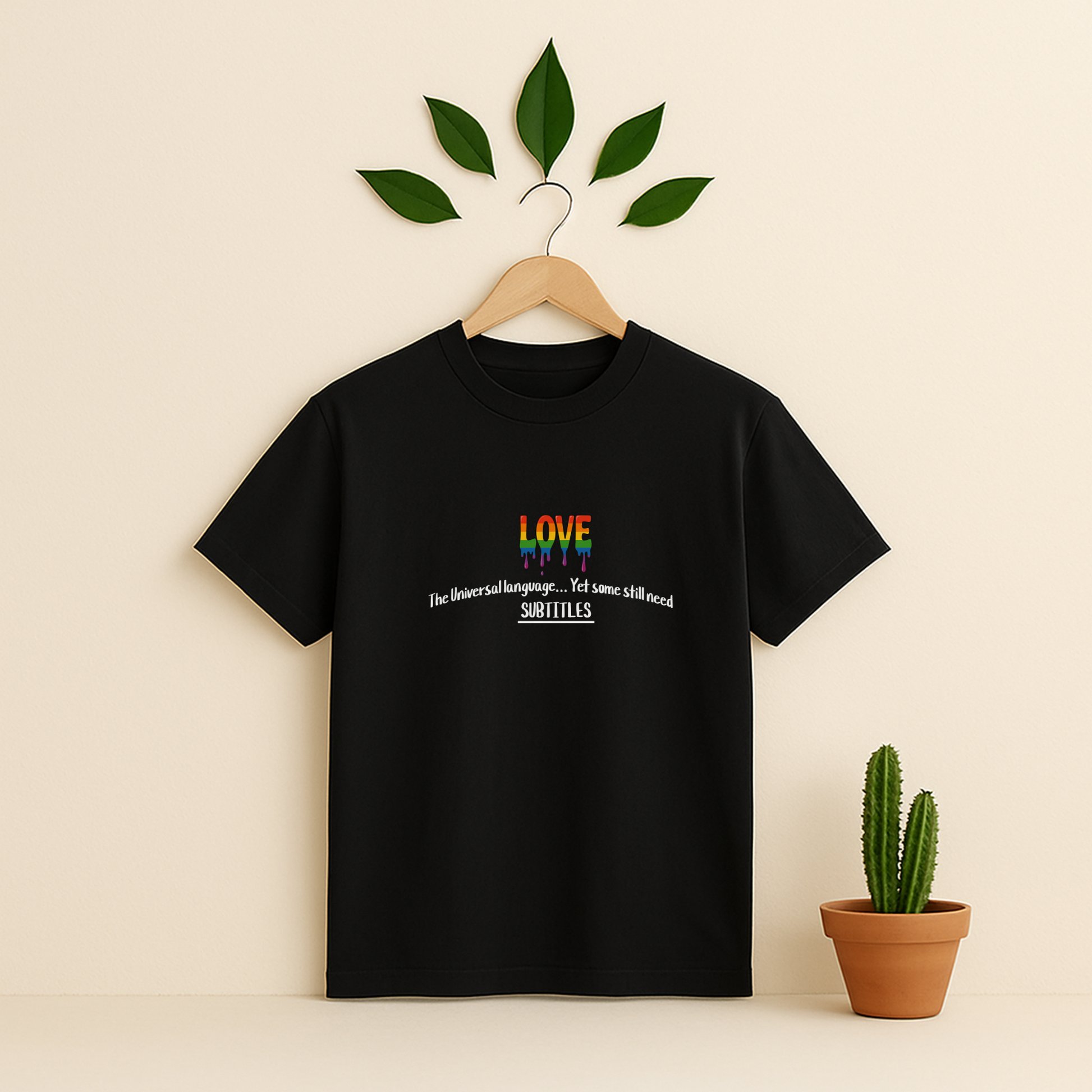 Black t-shirt with rainbow design and text on a wooden hanger against a beige background with a cactus plant.