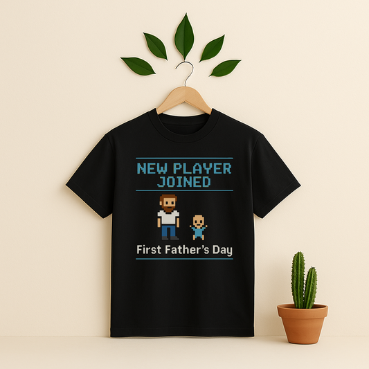 Black t-shirt with 'New Player Joined First Father's Day' graphic text and pixel art on a wooden hanger against a beige background with a cactus plant.