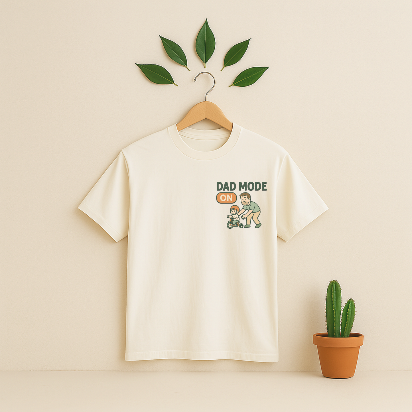 Ivory t-shirt with 'Dad Mode On' graphic on a wooden hanger against a beige background with a cactus plant.