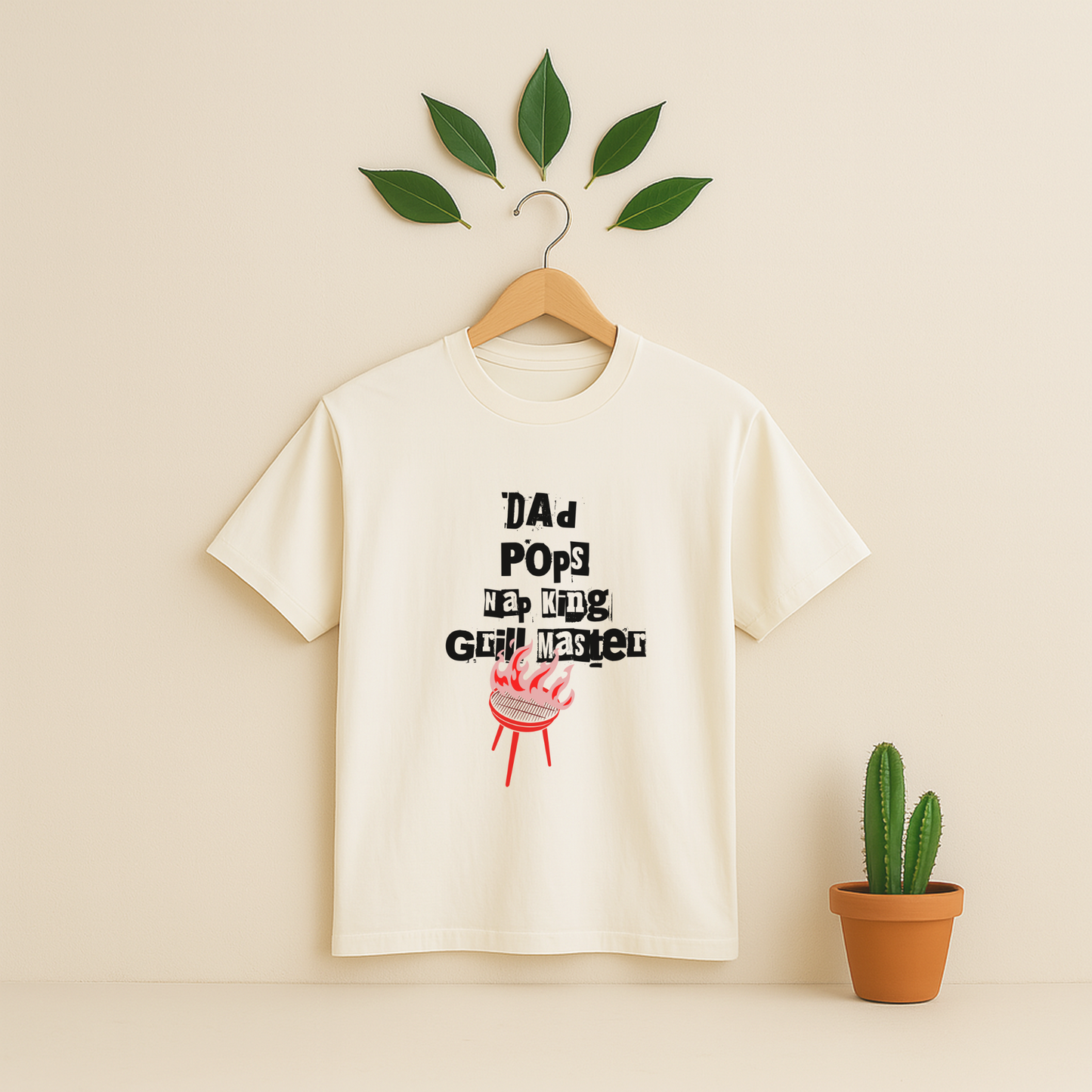 Ivory t-shirt with 'Dad Pops Grill Master' graphic on a wooden hanger against a beige background with a cactus plant.