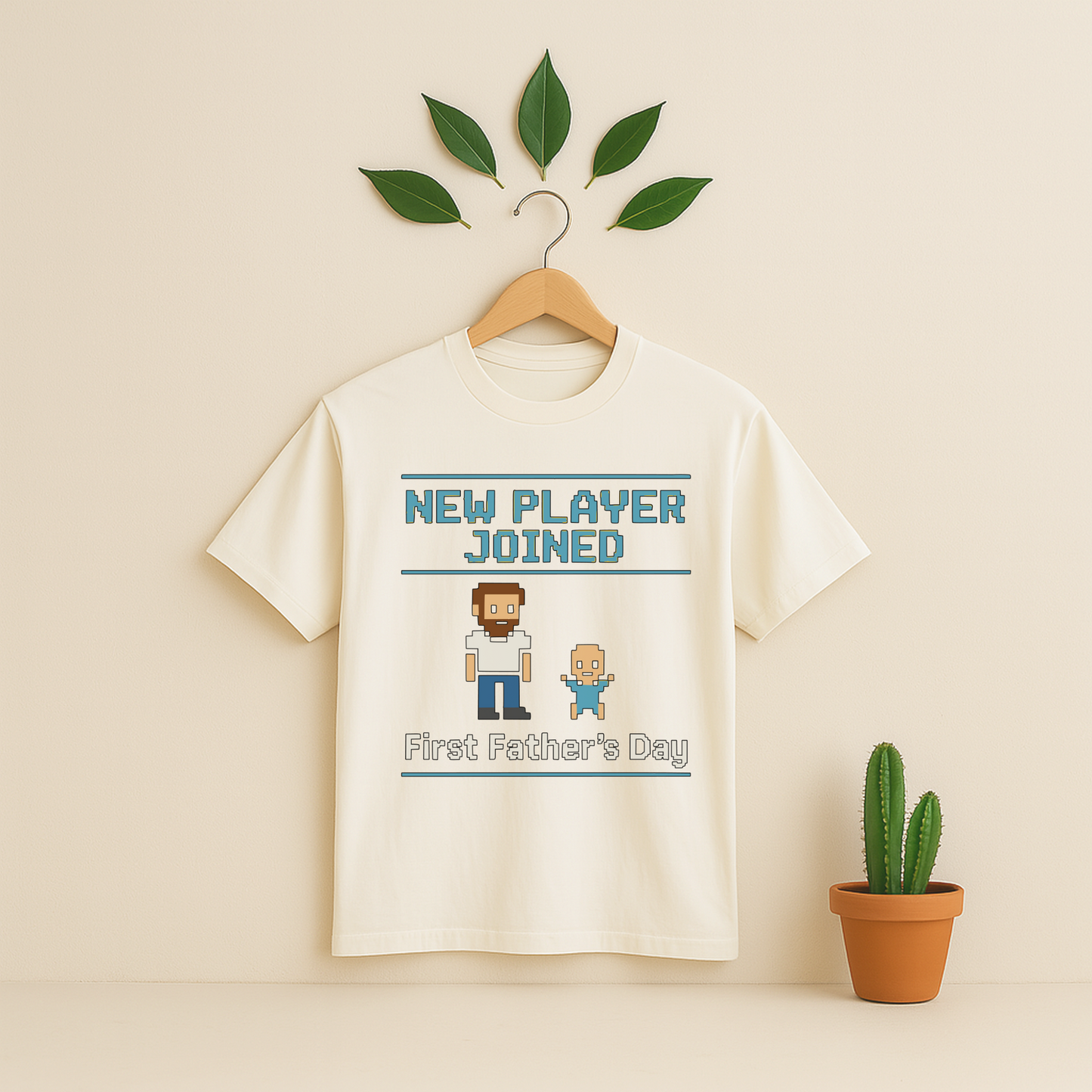 Ivory t-shirt with 'New Player Joined First Father's Day' graphic text and pixel art on a wooden hanger against a beige background with a cactus plant.