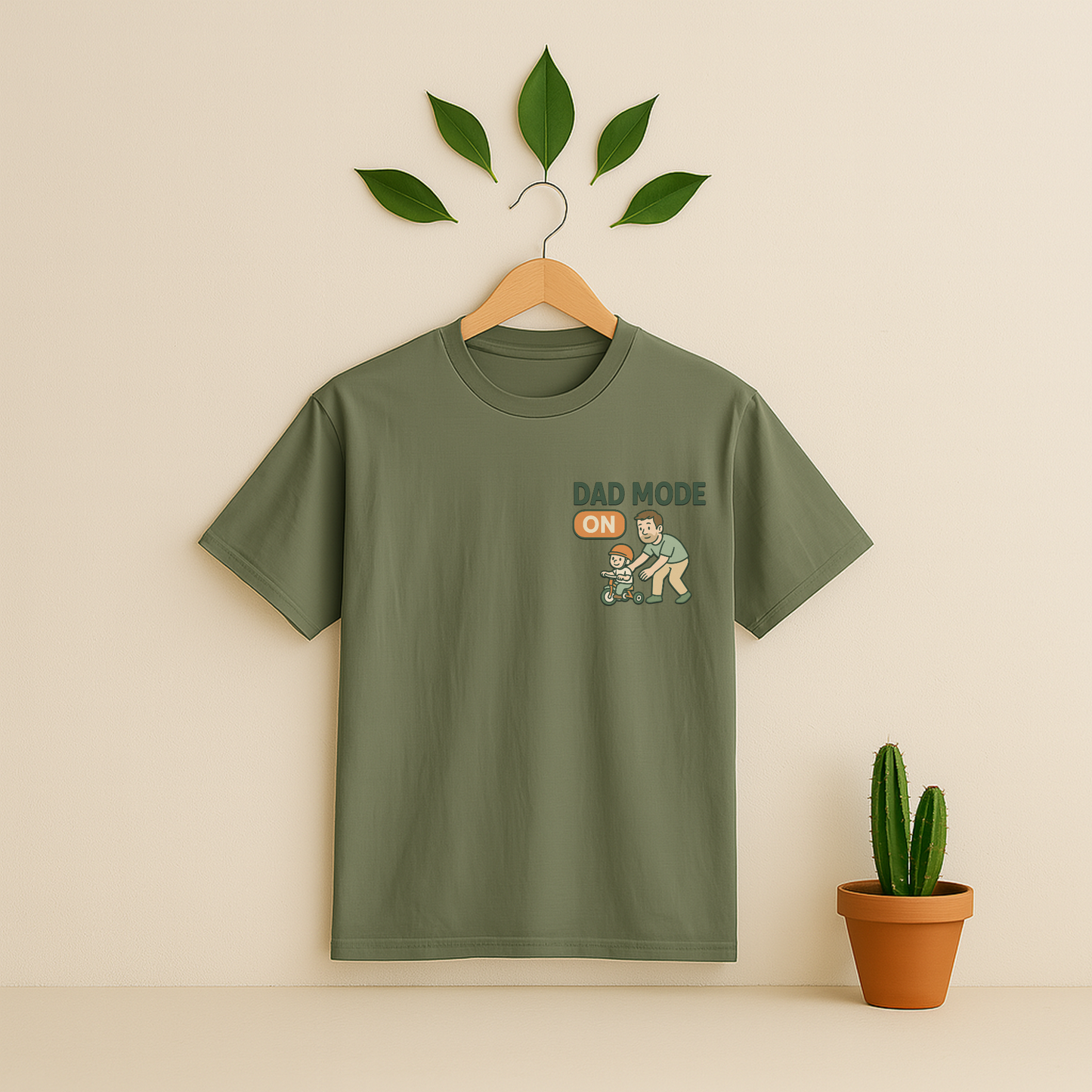 Moss t-shirt with 'Dad Mode On' graphic on a wooden hanger against a beige background with a cactus plant.