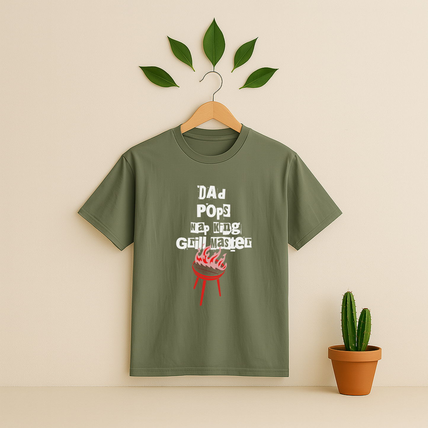 Moss t-shirt with 'Dad Pops Grill Master' graphic on a wooden hanger against a beige background with a cactus plant.