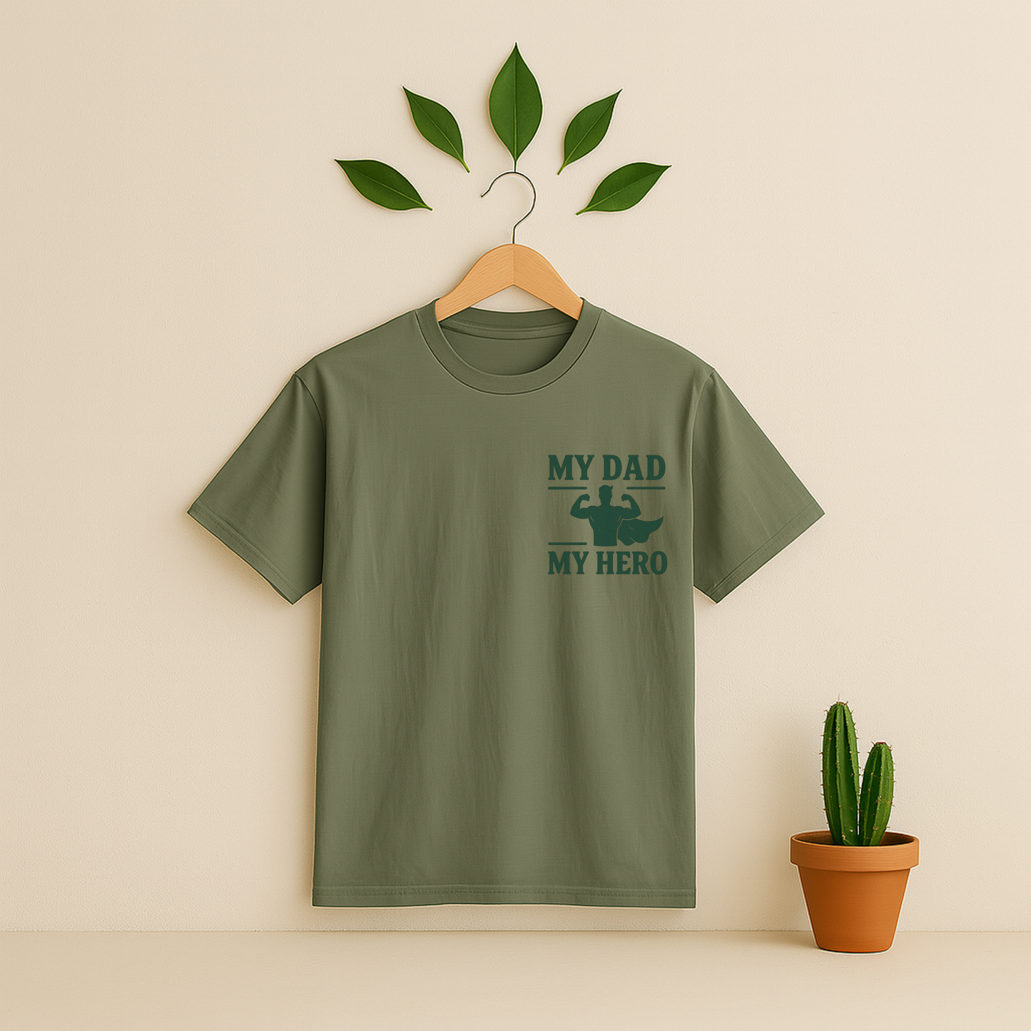 Moss t-shirt with 'My Dad My Hero' graphic on a wooden hanger against a beige background with a cactus plant.