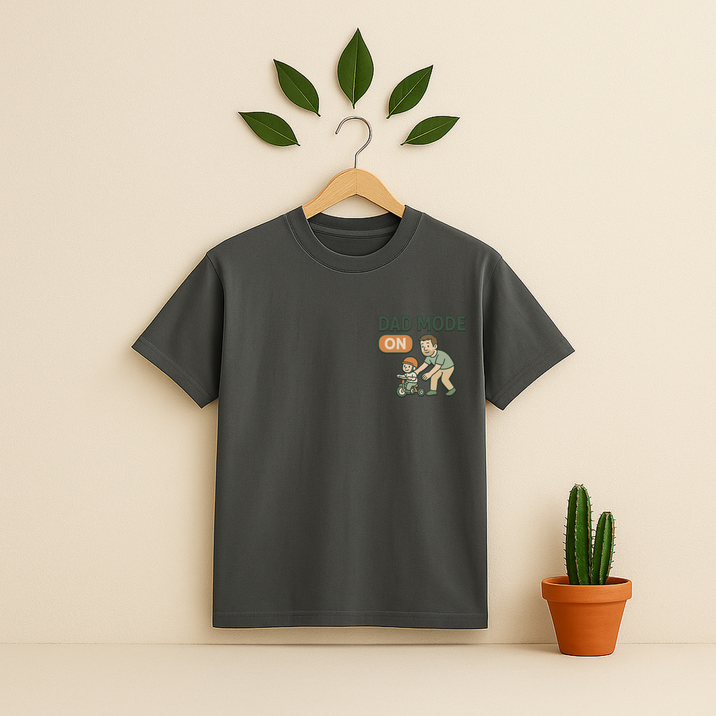 Pepper t-shirt with 'Dad Mode On' graphic on a wooden hanger against a beige background with a cactus plant.