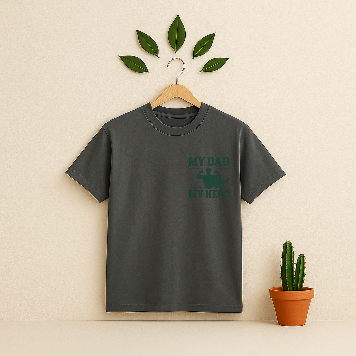 Pepper t-shirt with 'My Dad My Hero' graphic on a wooden hanger against a beige background with a cactus plant.
