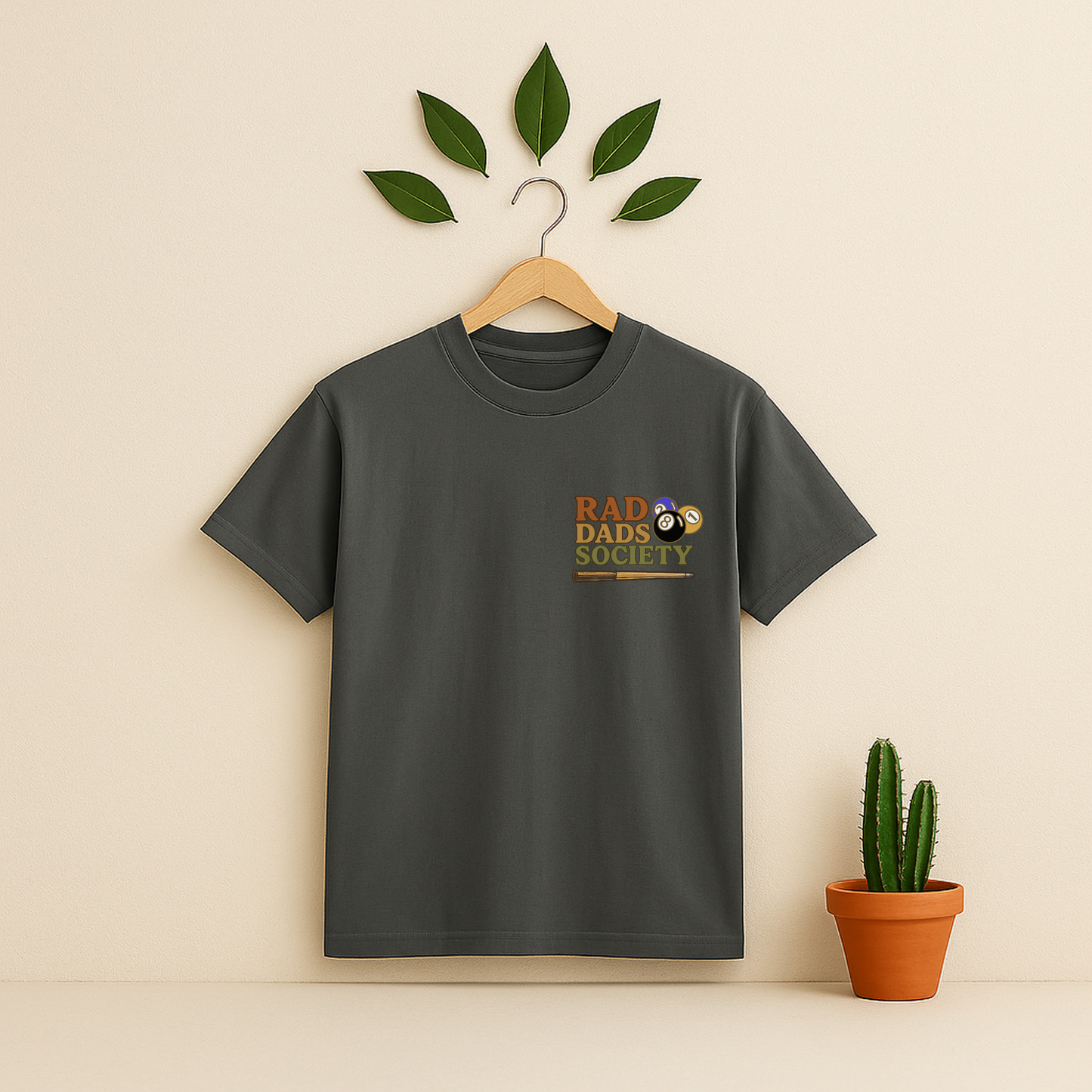 Pepper t-shirt with 'Rad Dads Society' graphic on a wooden hanger against a beige background with a cactus plant.
