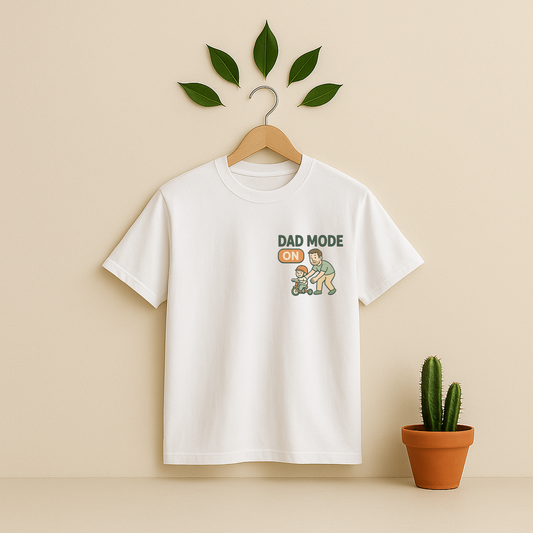 White t-shirt with 'Dad Mode On' graphic on a wooden hanger against a beige background with a cactus plant.