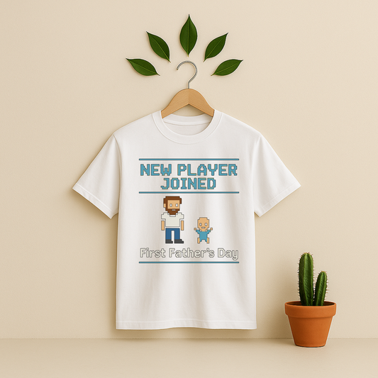 White t-shirt with 'New Player Joined First Father's Day' graphic text and pixel art on a wooden hanger against a beige background with a cactus plant.