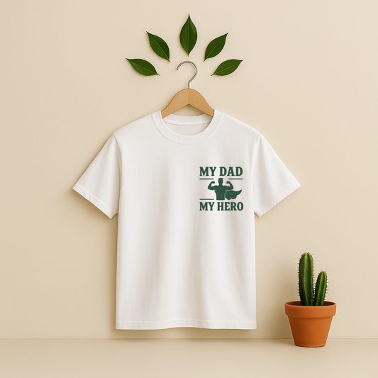 White t-shirt with 'My Dad My Hero' graphic on a wooden hanger against a beige background with a cactus plant.