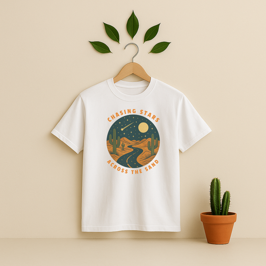 White t-shirt with 'Chasing Stars Across the Sand' graphic on a wooden hanger against a beige background with a cactus plant.