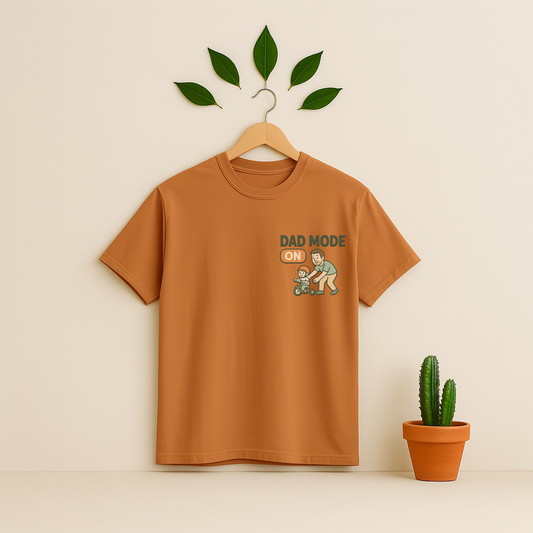 Yam t-shirt with 'Dad Mode On' graphic on a wooden hanger against a beige background with a cactus plant.