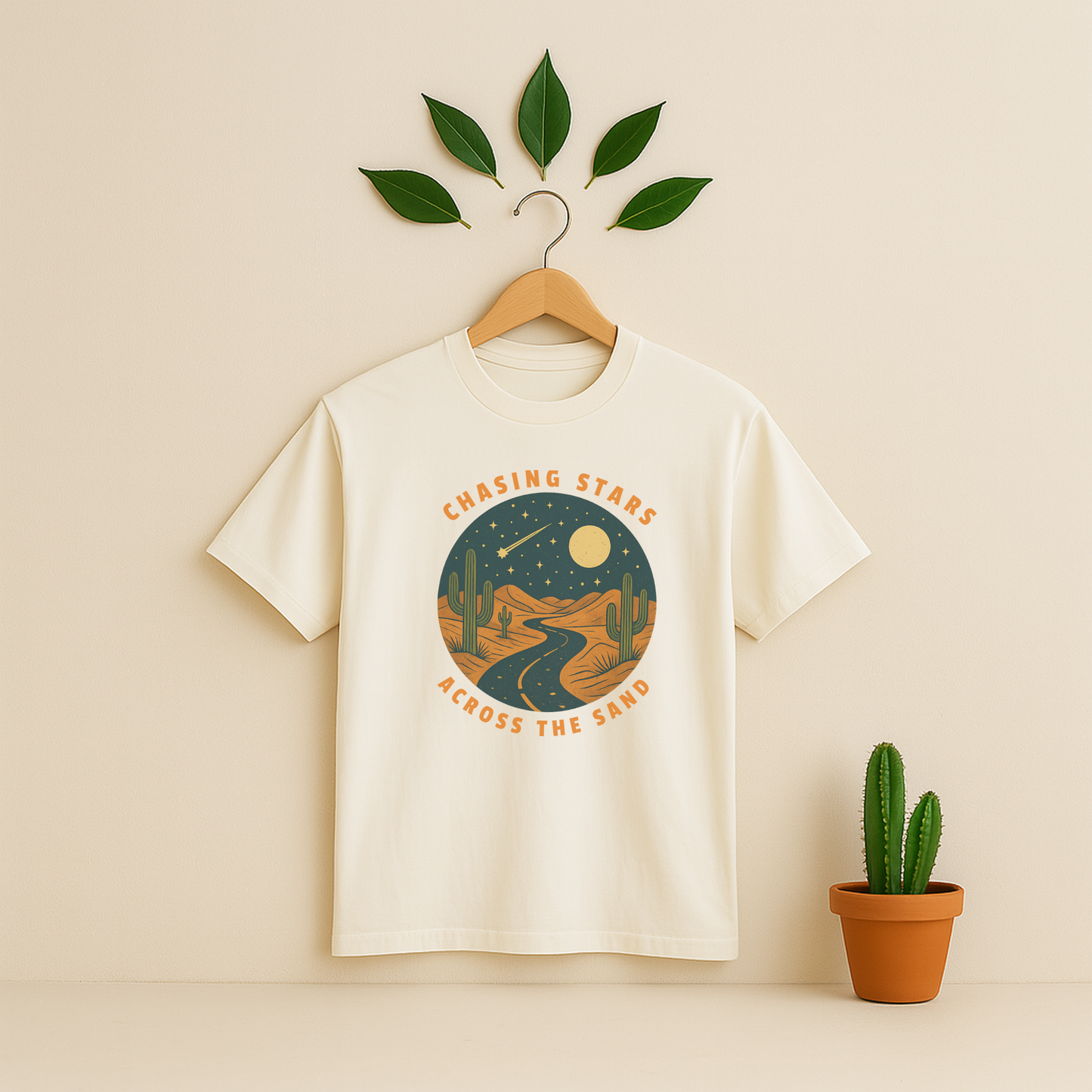 Ivory t-shirt with 'Chasing Stars Across the Sand' graphic on a wooden hanger against a beige background with a cactus plant.