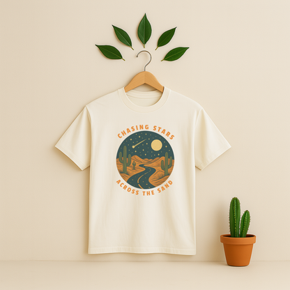 Ivory t-shirt with 'Chasing Stars Across the Sand' graphic on a wooden hanger against a beige background with a cactus plant.