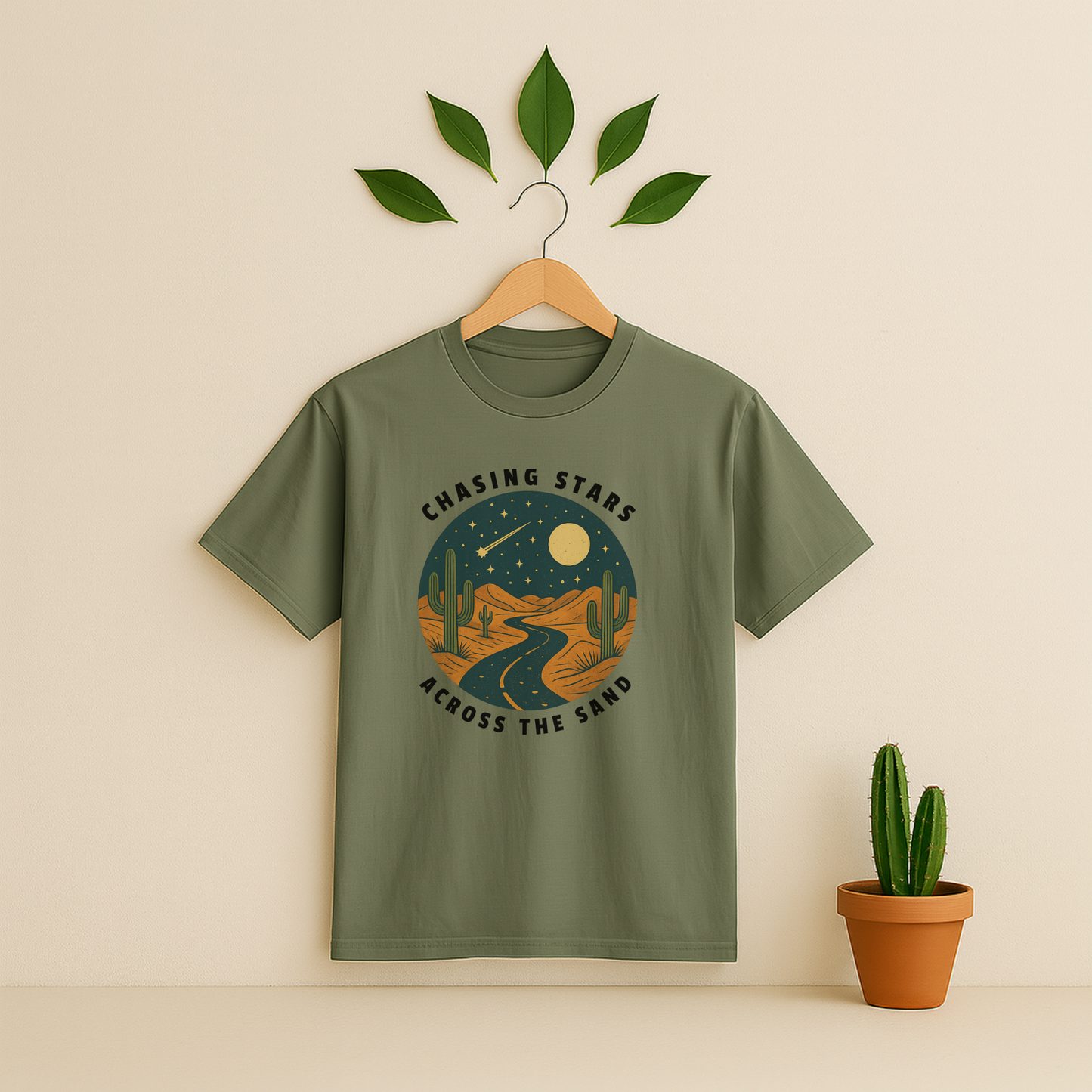 Moss t-shirt with 'Chasing Stars Across the Sand' graphic on a wooden hanger against a beige background with a cactus plant.