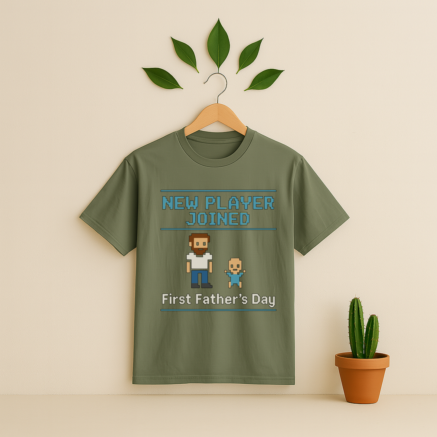 Moss t-shirt with 'New Player Joined First Father's Day' graphic text and pixel art on a wooden hanger against a beige background with a cactus plant.