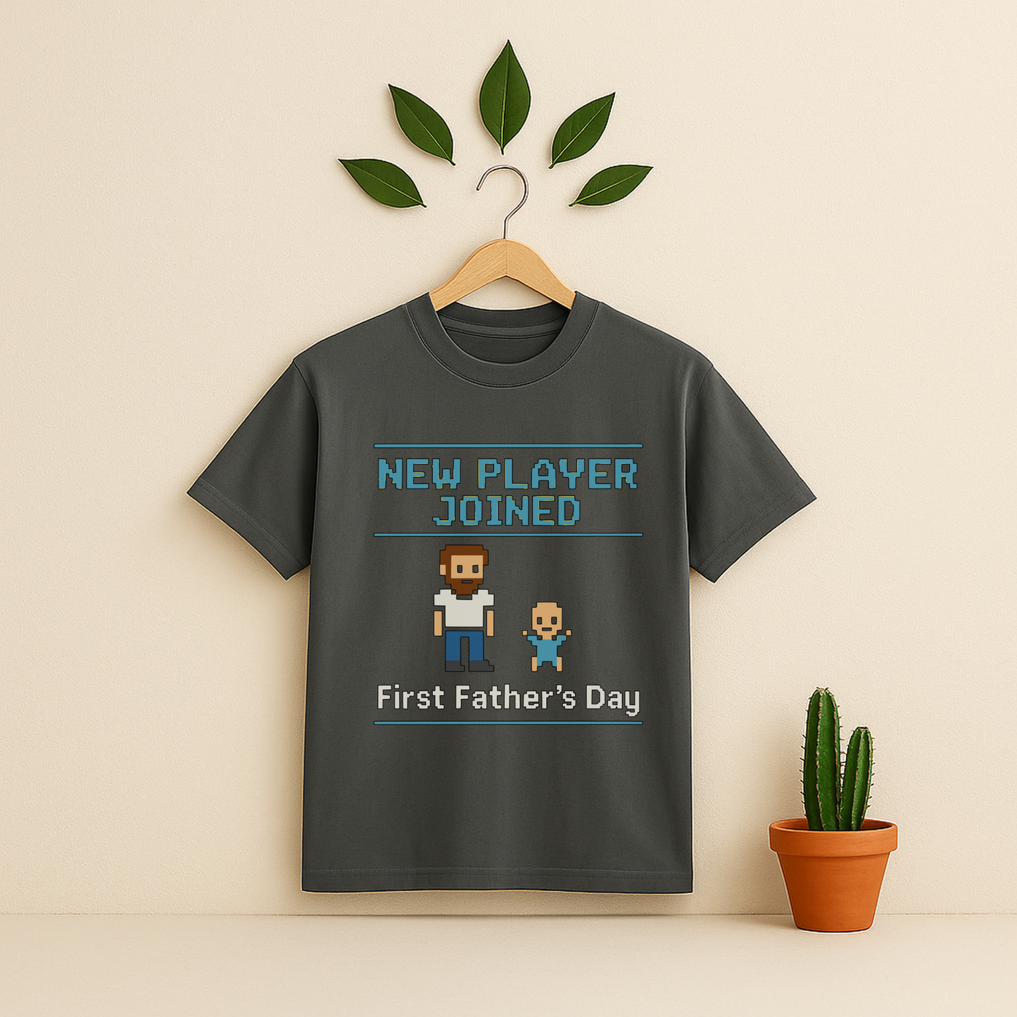 Pepper t-shirt with 'New Player Joined First Father's Day' graphic text and pixel art on a wooden hanger against a beige background with a cactus plant.
